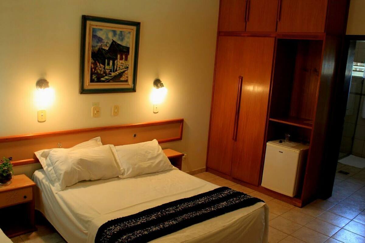 Un dormitorio con cama, lámparas de noche, mesita de noche, armario de madera y un refrigerador pequeño.