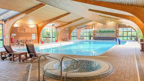 Une piscine intérieure avec un jacuzzi, des chaises en bois et un mur de briques orné d’une fresque.
