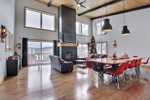 A spacious living area with a fireplace, a dining table with red chairs, and a large window overlooking a snowy landscape. - L’Ascension | 4-Season Spa & Mountain View (Saint-Philémon)