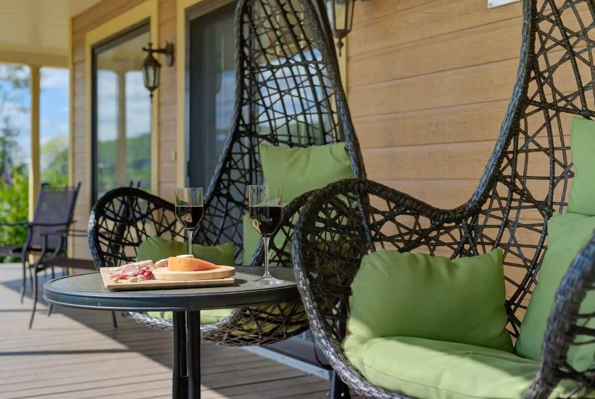 Una silla de mimbre con cojín verde sobre una terraza de madera, una mesa con un plato de comida y una copa de vino tinto, y vistas a la vegetación al fondo.