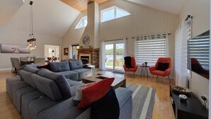 Een ruime woonkamer met een grote grijze hoekbank, een open haard en een eethoek met een tafel en stoelen.