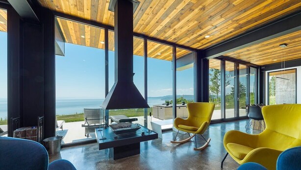 A modern interior with a large fireplace, a dining area, and a view of the outdoors. - L’Eaurizon | Private Spa & St. Lawrence View (La Malbaie)