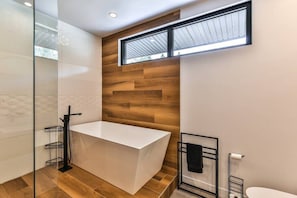 A modern bathroom with a white bathtub, wooden wall paneling, a glass shower enclosure, and a window above the bathtub.