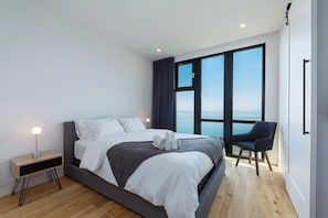 A modern bedroom with a large bed, a bedside table, a chair, and a view of the sea through a large window.