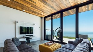 A modern living room with a large TV, comfortable seating, and a view of the sea. - L’Eaurizon | Private Spa & St. Lawrence View (La Malbaie)