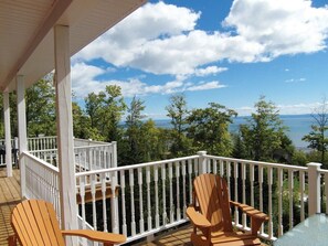 A covered deck with white railings, two wooden chairs, and a view of a lake and trees.