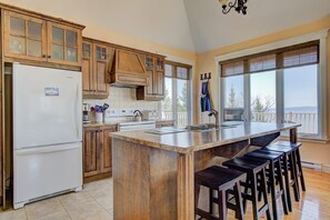 Une cuisine avec des meubles en bois, un réfrigérateur blanc, un grand îlot central avec des tabourets et une vue par les fenêtres.