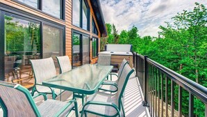 A modern outdoor deck with a glass table and chairs, a barbecue grill, and a view of a forested area.
