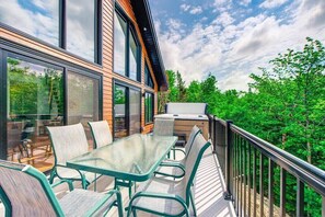A modern outdoor deck with a glass table and chairs, a barbecue grill, and a view of a forested area.