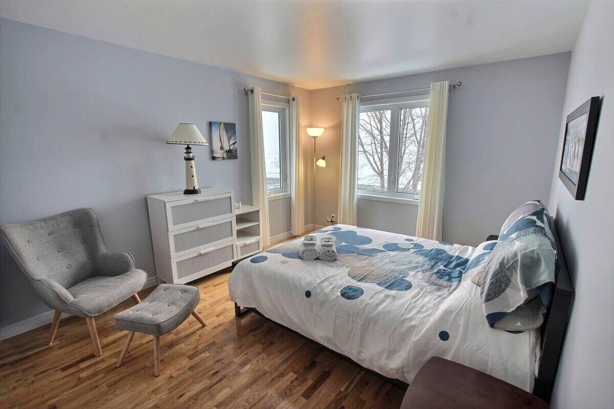 Un dormitorio con cama, una silla, una mesita de noche, una lámpara y una ventana.