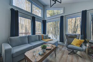 A modern living room with a grey sofa, a wooden coffee table, and a grey armchair with a yellow cushion. Large windows offer a view of trees outside. - Le Baikal | Nature, Trails & EV Charger (Pont-Rouge)