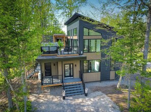 Ett modernt hus i två våningar med en balkong, stora fönster och en grusuppfart som omges av träd.