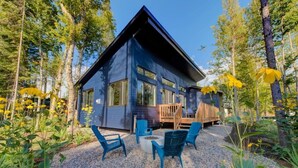 A modern cabin with a deck, surrounded by trees and yellow flowers.