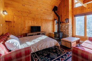 A wooden cabin interior with a bed, a television, a stone fireplace, and a window with a view of trees. - Boisé du Midi 66C | Hiking & Ski Escape (Saint-Philémon)