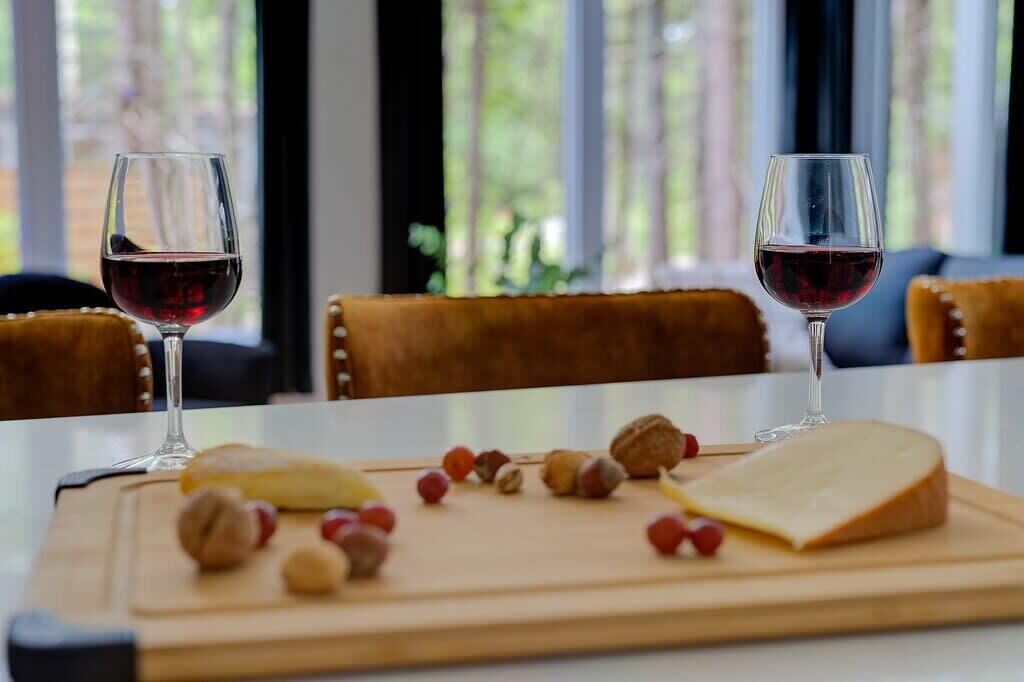 Una mesa con dos copas de vino tinto, una tabla de quesos con nueces y frutas, y una tabla de cortar de madera.