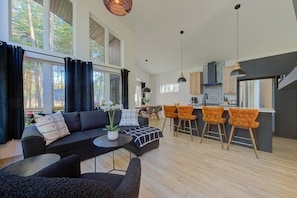 A modern living room with a sofa, ottoman, and chairs, a kitchen with bar stools, and large windows with curtains.