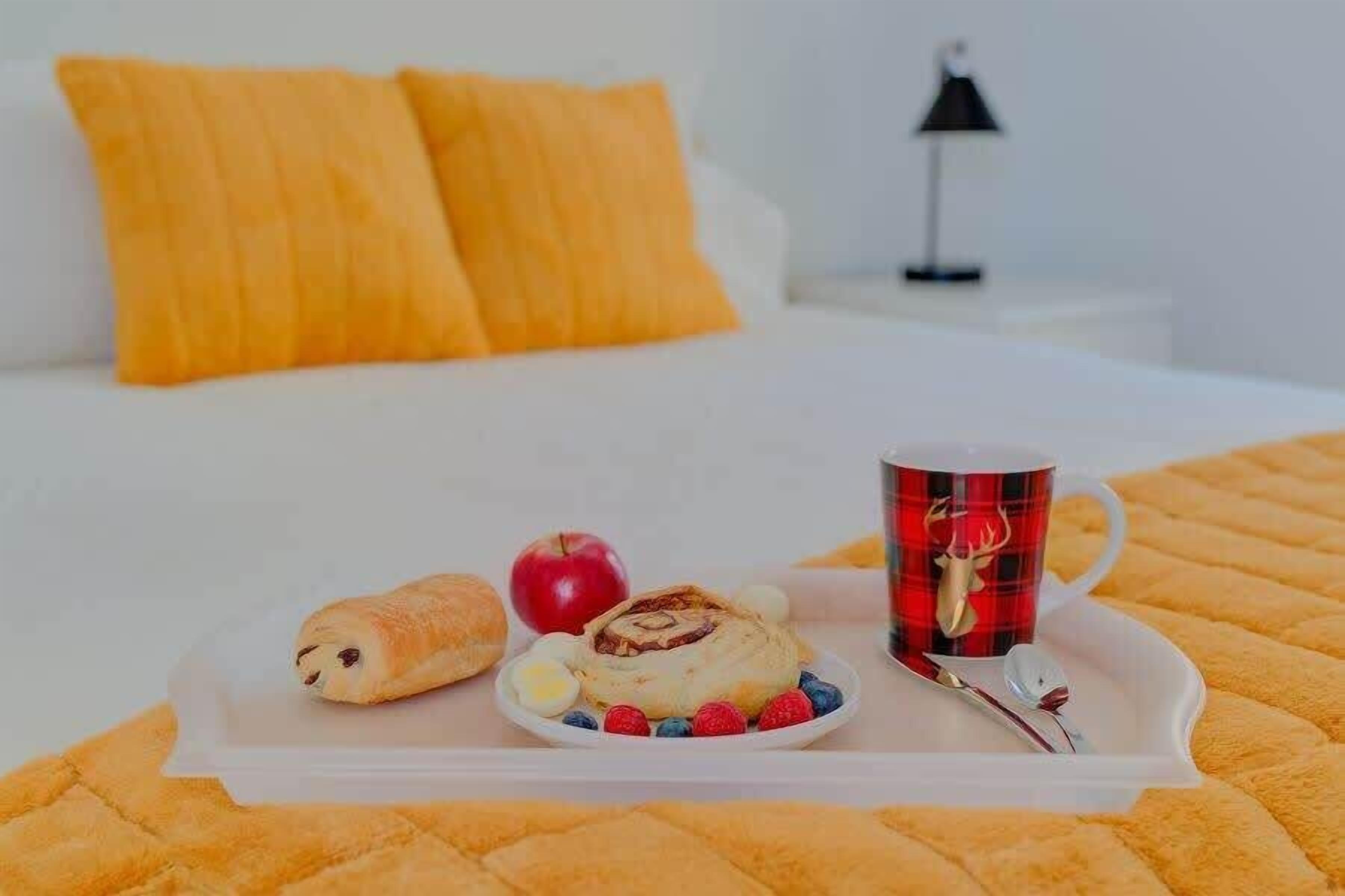 A bed with a white bedspread, a tray with breakfast items including a croissant, a cinnamon roll, an apple, and berries, and a mug with a deer design.