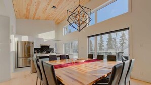 Une cuisine moderne avec un réfrigérateur en acier inoxydable, un coin repas doté d’une grande table en bois et de chaises noires, et de grandes fenêtres offrant une vue sur les arbres.
