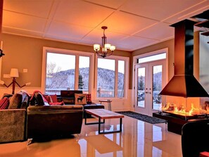 A spacious living room with a fireplace, large windows, and a chandelier.