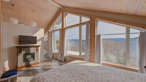 A bedroom with a stone fireplace, a television, and large windows offering a view of the outdoors. - Pourquoi Pas! - Hot tub and Ski, Mountain view (Saint-Philémon)