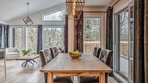 Un espace repas comprenant une table en bois, des chaises en cuir, un luminaire suspendu et de grandes fenêtres donnant sur la forêt.