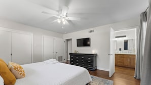 Une chambre à coucher avec un lit, un ventilateur de plafond, une commode, un téléviseur et une salle de bain visible à travers une porte ouverte.
