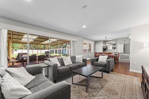 A spacious living room with a large sofa set, a coffee table, and a patterned rug. There is a dining area with a table and chairs, and a kitchen in the background. - Cozy+Warm for Rest or Play PingPong+Fire pit+Grill (Baton Rouge)