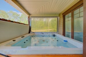 A hot tub with a view of a forest, a covered patio, and a window with blinds.