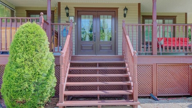 Una escalera de madera que conduce a un par de puertas dobles, un árbol pequeño y una silla roja en un porche.