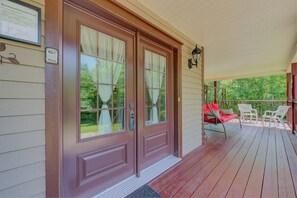 A wooden front door with glass panels, a porch with a red bench and white chairs, and a view of greenery.