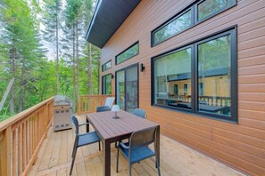 A wooden deck with a table and chairs, large windows, and a view of a forest.