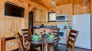 A wooden cabin interior with a dining table set for four, a kitchen area with a microwave and refrigerator, and a television mounted on the wall. - Le Huard - Lakefront & Private Dock (Sayabec)