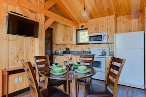 Houten chalet interieur met een eettafel voor vier personen, een keuken met een magnetron en koelkast, en een televisie aan de muur bevestigd.