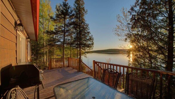 A wooden deck with a table and chairs, overlooking a lake with trees and a building in the background. - Le Huard - Lakefront & Private Dock (Sayabec)