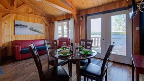 A wooden cabin interior with a dining table set for two, a red sofa, and a painting of a boat on the wall. - Le Huard - Lakefront & Private Dock (Sayabec)