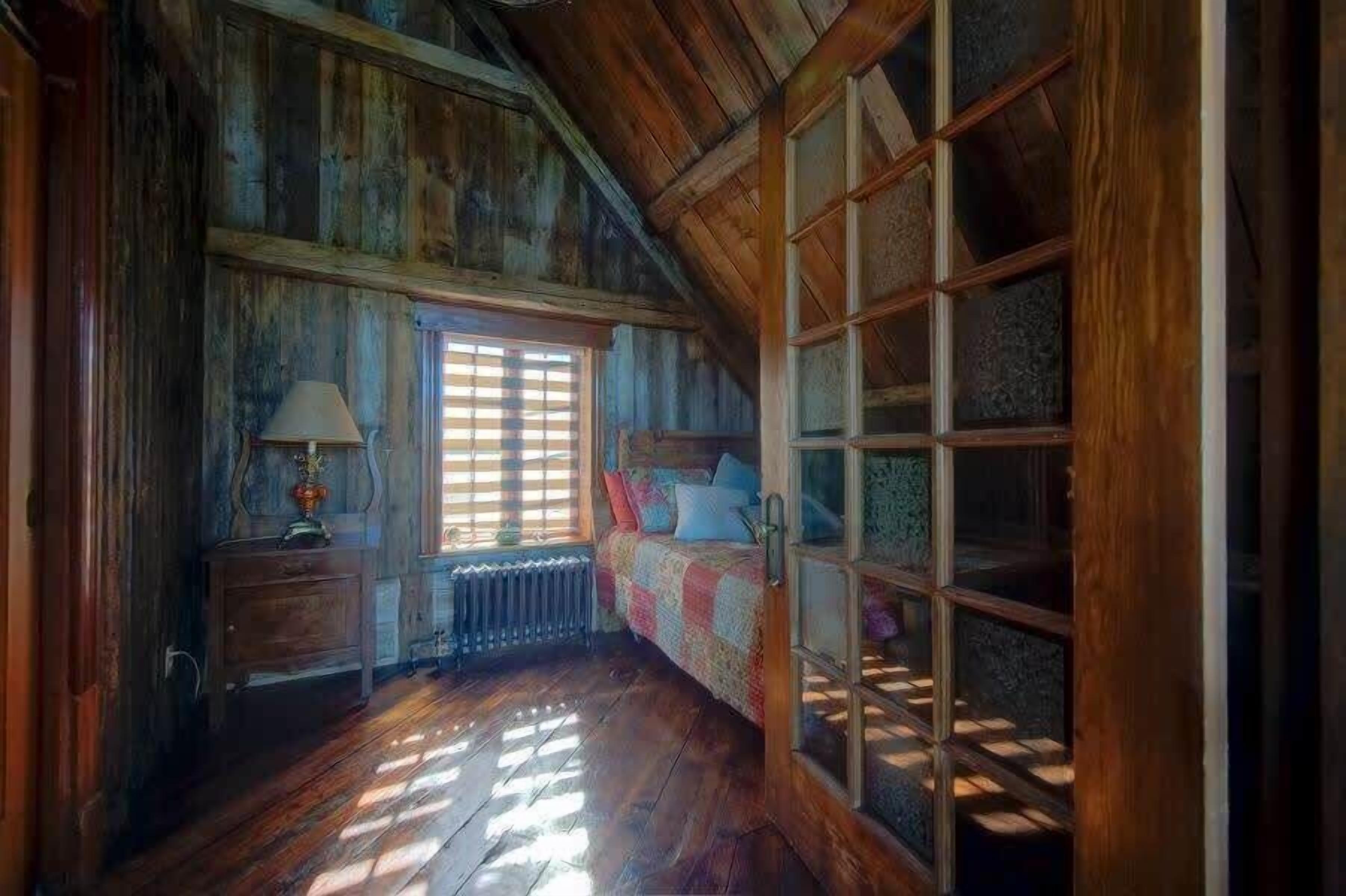 A room with wooden walls, a window with blinds, a bed with a quilt, a nightstand with a lamp, and a radiator.