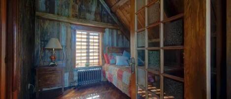 A room with wooden walls, a window with blinds, a bed with a quilt, a nightstand with a lamp, and a radiator.
