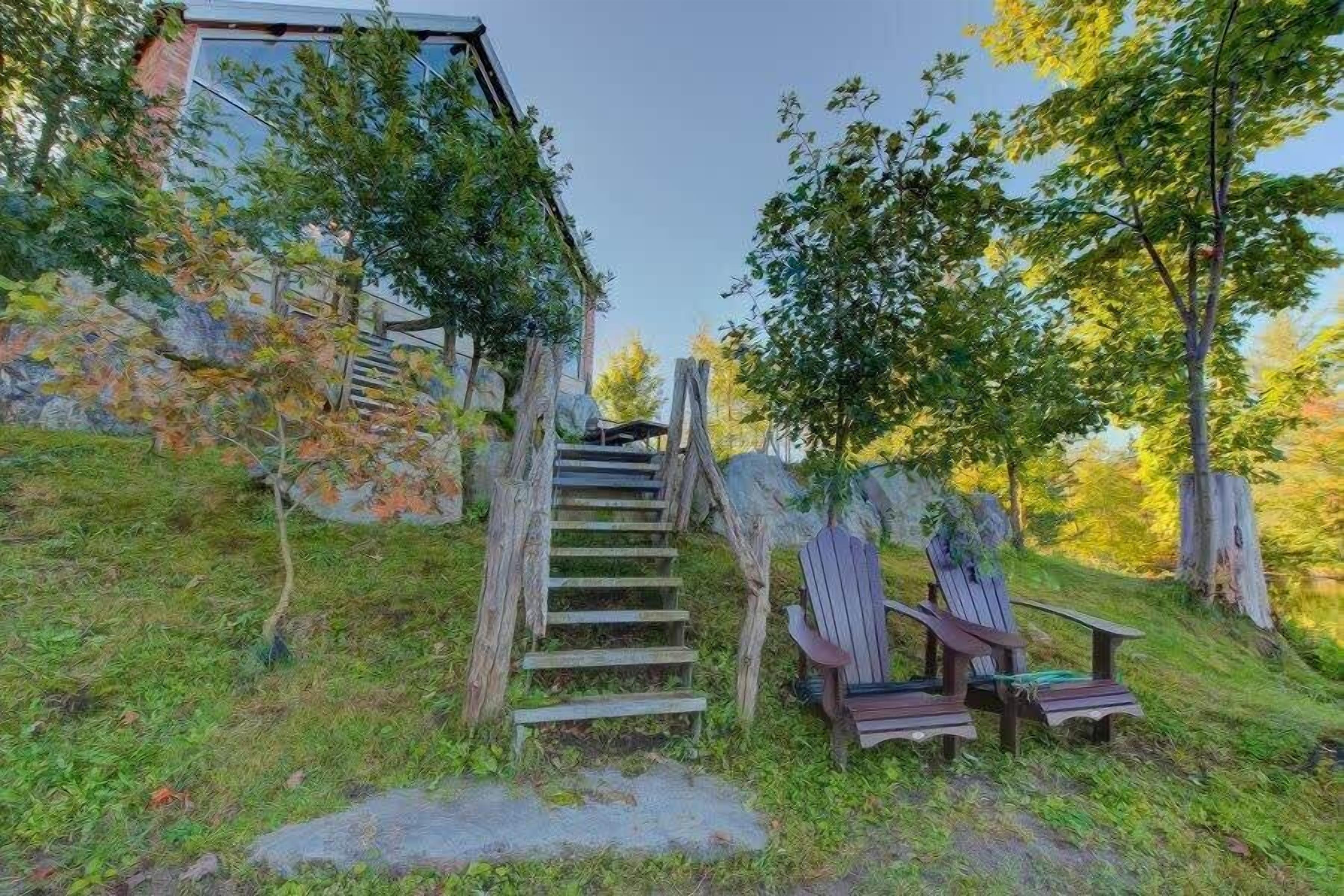 A wooden staircase leading to a building, surrounded by trees and two Adirondack chairs.