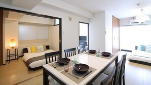 A modern hotel room with a bed, a dining table set with plates and glasses, a bedside lamp, and a view of the city through large windows.