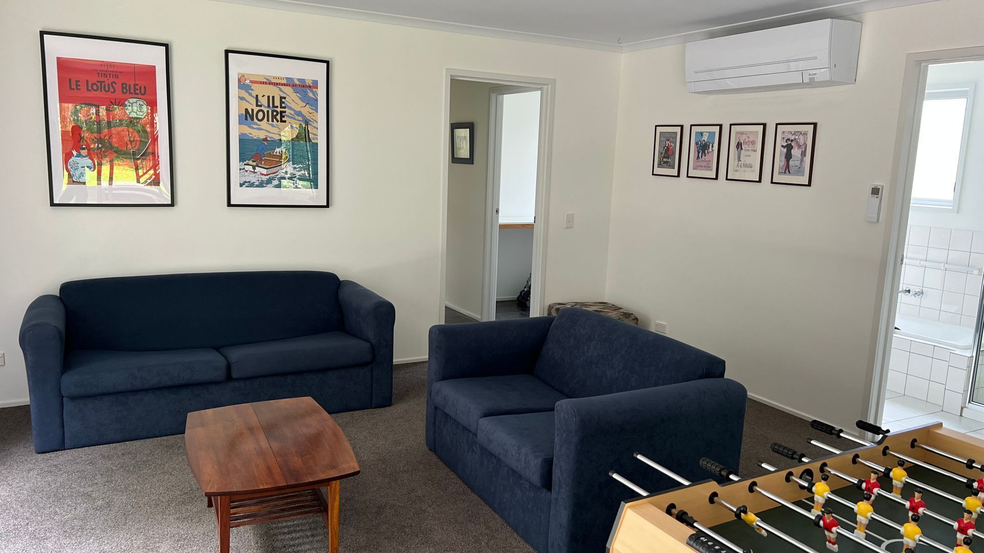 A living room with a dark blue sofa, a wooden coffee table, and a foosball table. There are two framed posters on the wall and an air conditioning unit above.