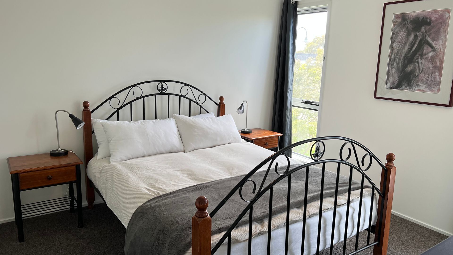 A bed with a metal frame, two bedside tables, a window with a view, and a framed artwork on the wall.