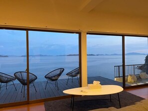 A modern room with large windows overlooking a body of water, a wooden deck, and a round table with chairs. - Exclusive Seaside Escape in Ocean House (Kure)