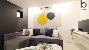 A modern living room with a grey sectional sofa, a white coffee table with books and a decorative bowl, a flat-screen TV on a white stand, and a wall-mounted air conditioner. - b hotel 102 Central Inner-Side Comfort Studio (Hiroshima City)