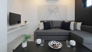 Un salon moderne avec un canapé gris, des coussins blancs et noirs, une table basse noire sur laquelle se trouvent un bol blanc et des verres, un téléviseur à écran plat posé sur une étagère blanche et un climatiseur fixé au mur.