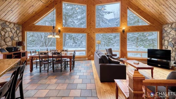 Amplia sala de estar con pared de piedra, techo de madera y ventanas grandes que dan a árboles cubiertos de nieve.