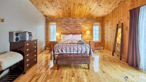 Un dormitorio acogedor con techo de madera, pared de ladrillo, una cama con cabecera, lámparas de noche, cómoda y un banco.