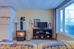A cozy room with a wood-burning stove, a television on a wooden stand, a window with a view, and a bed with patterned bedding.