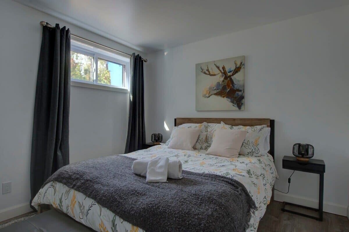 A bedroom with a bed, a window with curtains, a wall-mounted artwork, and a bedside table with a lamp.