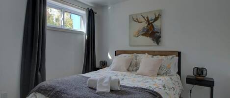 A bedroom with a bed, a window with curtains, a wall-mounted artwork, and a bedside table with a lamp.