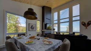 Spiseplads med et bord dækket til et måltid, en sofa og store vinduer med udsigt til træer.
